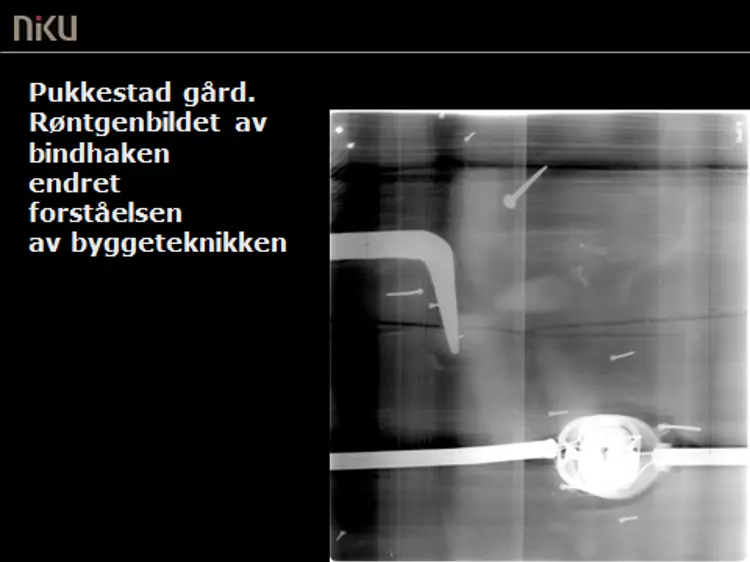 Røntgenbilde av et utsnitt av laftet og tosidig panelt yttervegg på Pukkestad gård, Sandefjord.  Bindhaken (pil) som ble oppdaget ved hjelp av røntgenundesøkelsen, vakte stor forbauselse. Ytterliger undersøkelser viste at bindhaken ligger nedfelt i laftestokken, slik at den kun ville blitt oppdaget dersom bygningen ble revet. Samtlige stokker på denne ytterveggen var, på grunn av veggens lengde,  skjøtet ved hjelp av bindhaker. Dette er en svært uvanlig bygningsteknikk. Foto: NIKU