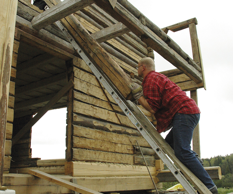 Kraftkaren Kåre Brynjulvsrud skyver her opp en av stokkene til andre etasje, mens jeg, Tor harald Frøyset, drar den opp etter tauet. Ved å legge en planke på stigen på denne måten, klarte vi fint å få opp tunge stokker. Foto: Norske Hjem