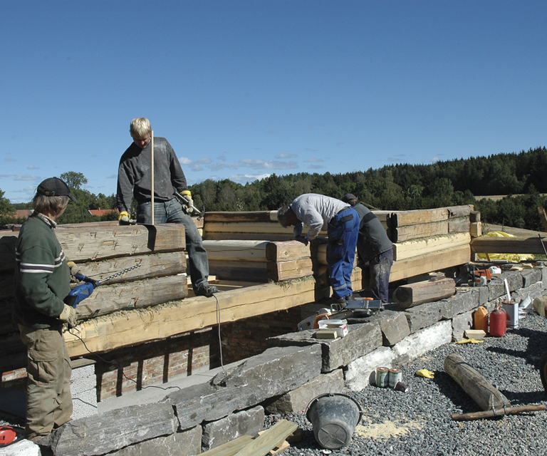 Her er vi godt i gang med å lafte opp de gamle stokkene. Det bores opp hull til treplugger/ dumlinger som hjelper til å holde tømmerveggene stabile. Foto: Norske Hjem